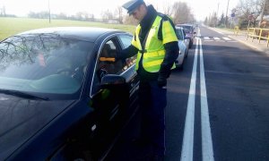Policjant stoi na jezdni przy samochodzie koloru czarnego. Przy użyciu alkomatu kontroluje kierowcę.