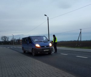 Na drodze stoi policjant podchodzi do niebieskiego samochodu typu bus trzymając w ręku alkomat.