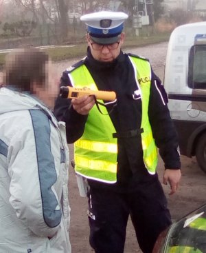 Policjant ruchu drogowego podczas przeprowadzenia badania stanu trzeźwości alkomatem. Po prawej stronie widoczny pojazd kierującego.