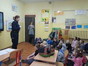 Wnętrze sali lekcyjnej. Z prawej strony st. sierż. Katarzyna Leszczyńska - Krawczyk. Z lewej  strony dzieci siedzące na dywanie. Pod ścianą stoi pani nauczycielka.