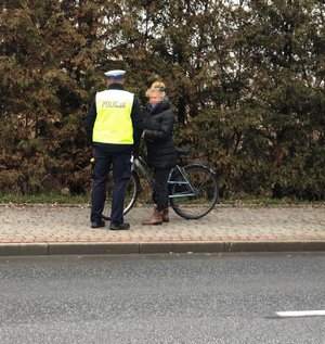 Na tle żywopłotu, na chodniku stoi policjant i rowerzysta, który jest legitymowany.