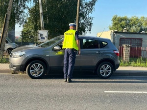 Policjant ruchu drogowego podczas kontroli pojazdu osobowego. Funkcjonariusz soi przy drzwiach od strony kierowcy. pojazd stoi na chodniku.