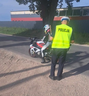 Policjant ruchu drogowego stoi na poboczu drogi. Przed nim na jezdni znajduje się motocykl, a na nim siedzi kierująca. W tle hala produkcyjna.