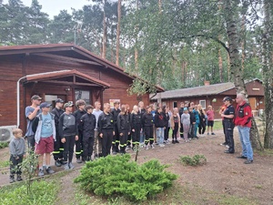 Przed drewnianymi kwaterami ZHP stoi w dwuszeregu młodzież OSP. Przed nimi policjantka wraz z opiekunami.