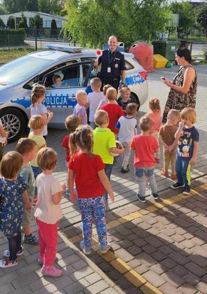 /policjant przed radiowozem. W ręku trzyma tarczę do zatrzymywania pojazdów i pokazuje ją dzieciom zebranym wokół niego.