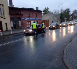 Na osi jezdni stoją policjanci drogówki w żółtych kamizelkach. Z prawej strony nadjeżdża kolumna pojazdów.