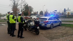 4 policjantów ruchu drogowego ubranych w żółte kamizelki stoi w rejonie cmentarza na ul. 700-lecia w Zninie. Przed nimi motocykle i radiowóz z włączonymi niebieskimi światłami.