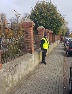 Policjant ubrany w żółtą kamizelkę, stoi przy ogrodzeniu cmentarza i obserwuje parkujące pojazdy.
