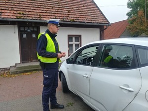 Policjant ruchu drogowego stoi przy białym samochodzie od strony kierowcy i prowadzi kontrolę drogową.