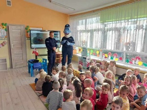 Klasa w przedszkolu. Asp. Dawid Rosół i Polfinek. Stoją przy rzutniku multimedialnym. Przed nimi na dywanie dzieci.