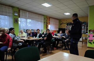 Dzielnicowy w świetlicy w Piechcinie opowiada seniorom zebranym przy stołach o oszustwach dokonywanych na osobach starszych.