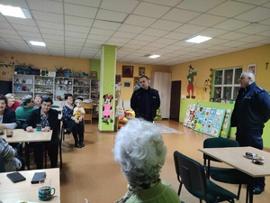 asp. Dawid Rosół i st. asp. Krzysztof Zientek podczas prelekcji dla seniorów. Z lewej strony starsze osoby siedzące przy stołach.