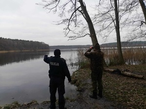 Teren przywodny. Policjant pokazuje ręką obszar jeziora. Funkcjonariusz Państwowej Straży Rybackiej patrzy przez lornetkę.